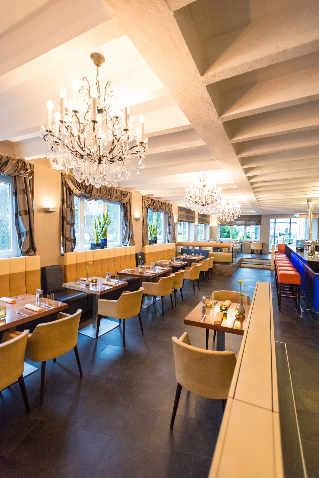 Elegantes Restaurant-Interieur mit Kronleuchtern, gedeckten Tischen, gepolsterten Stühlen und großen Fenstern, die natürliches Licht hereinlassen.