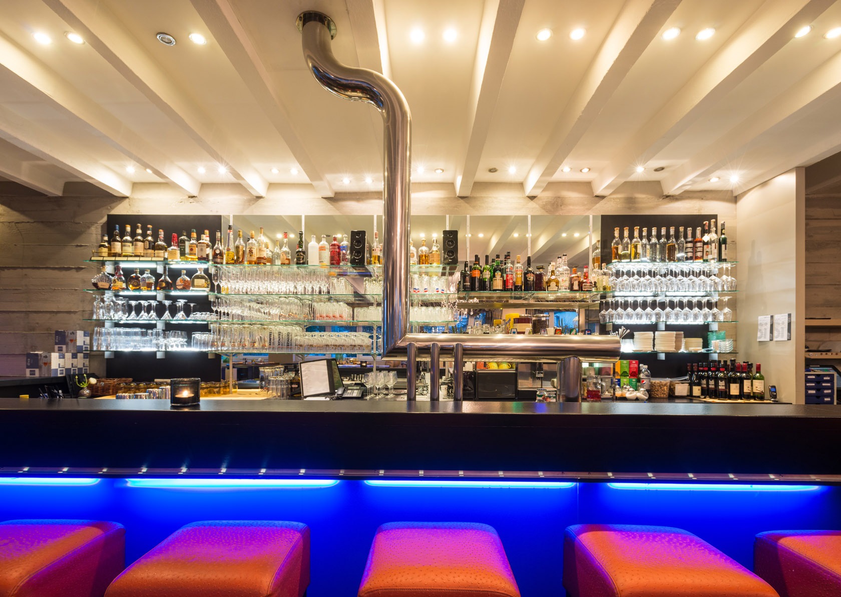 Moderne Bar mit beleuchteter blauer Theke, roten Barhockern und Regalen hinter der Bar voller Flaschen und Gläser.
