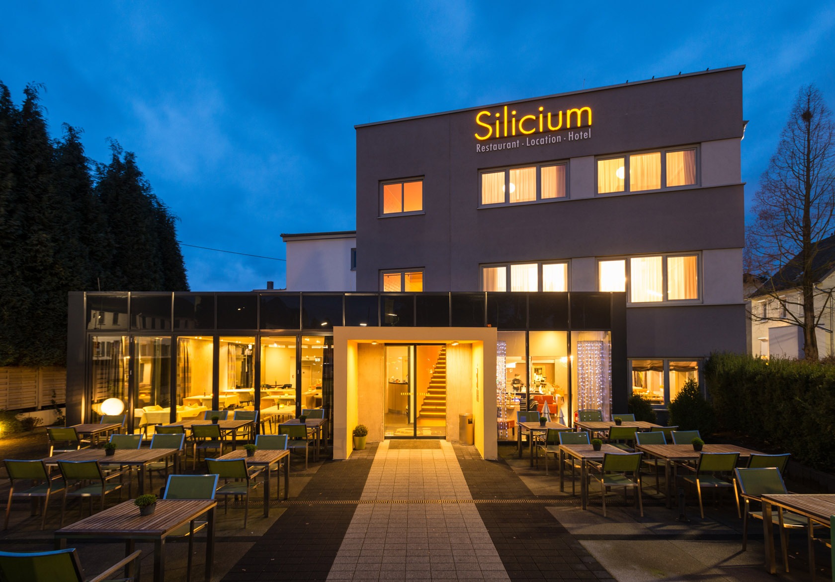 Modernes Hotel und Restaurant namens Silicium mit Sitzgelegenheiten im Freien, warm beleuchteten Fenstern und blauem Abendhimmel.