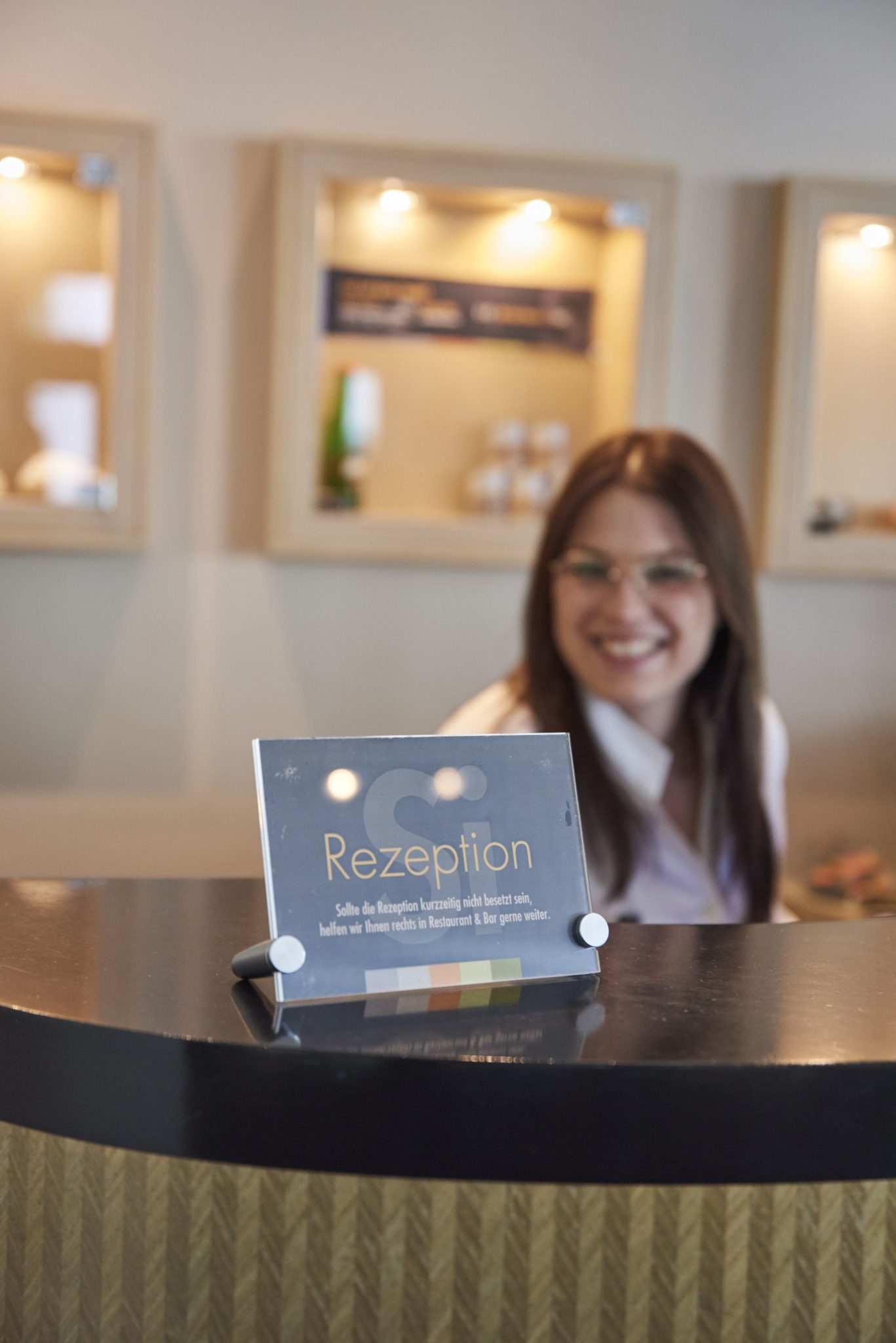 Eine lächelnde Frau steht hinter der Rezeption eines Hotels mit einem Schild, auf dem auf Deutsch „Rezeption“ steht.