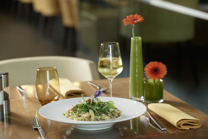 Ein Nudelgericht mit Gemüse und essbaren Blumen, serviert mit Weißwein, auf einem Holztisch mit gelben Servietten und orangefarbenen Blumen in Vasen.