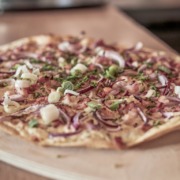 Eine Pizza mit dünnem Boden, belegt mit Zwiebeln, Frühlingszwiebeln, Speckstückchen und Schnittlauch, liegt auf einem Holzbrett.