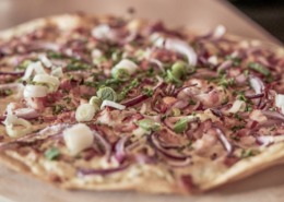 Eine Pizza mit dünnem Boden, belegt mit Zwiebeln, Frühlingszwiebeln, Speckstückchen und Schnittlauch, liegt auf einem Holzbrett.