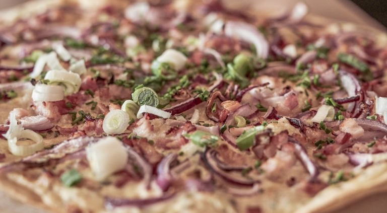 Eine Pizza mit dünnem Boden, belegt mit Zwiebeln, Frühlingszwiebeln, Speckstückchen und Schnittlauch, liegt auf einem Holzbrett.