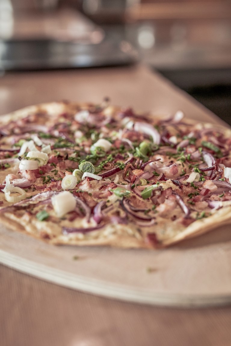 Eine Pizza mit dünnem Boden, belegt mit Zwiebeln, Frühlingszwiebeln, Speckstückchen und Schnittlauch, liegt auf einem Holzbrett.