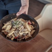 Eine Person serviert eine Schüssel Penne-Nudeln mit Pilzen, Tomaten und Microgreens auf einem Holztisch neben einem cremefarbenen Stuhl.