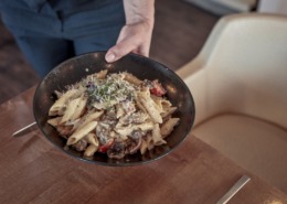 Eine Person serviert eine Schüssel Penne-Nudeln mit Pilzen, Tomaten und Microgreens auf einem Holztisch neben einem cremefarbenen Stuhl.