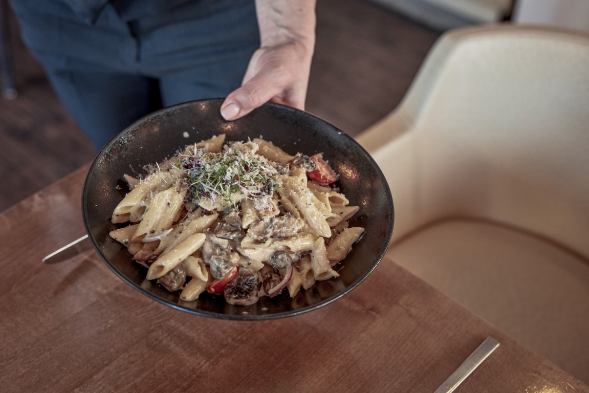 Eine Person serviert eine Schüssel Penne-Nudeln mit Pilzen, Tomaten und Microgreens auf einem Holztisch neben einem cremefarbenen Stuhl.