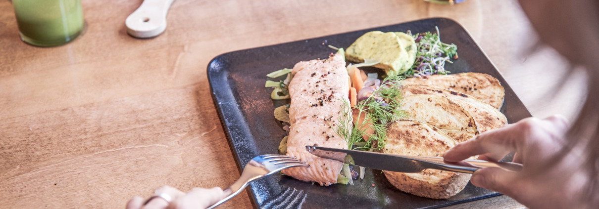 Person, die an einem Holztisch sitzt und gegrillten Lachs auf einem schwarzen Teller mit Toast, Gemüse und einem grünen Dip schneidet.