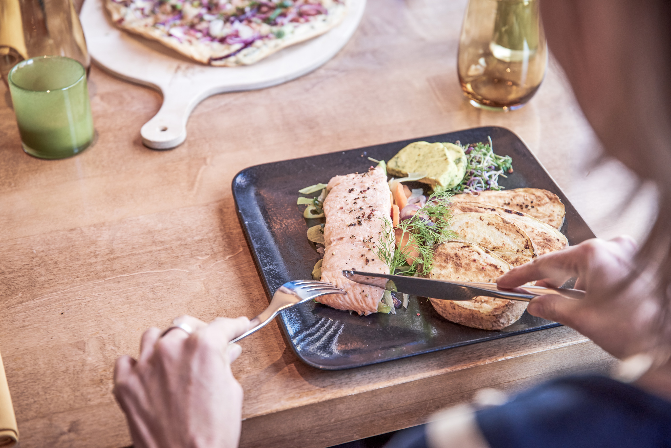 _BR15499 2_filter Person, die an einem Holztisch sitzt und gegrillten Lachs auf einem schwarzen Teller mit Toast, Gemüse und einem grünen Dip schneidet.