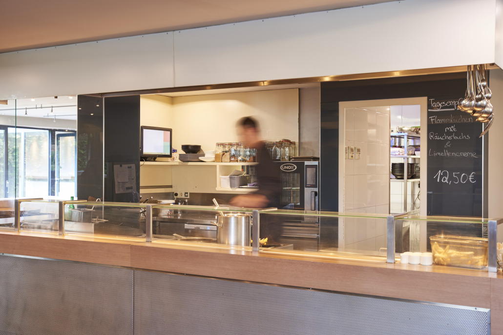Eine Person arbeitet hinter einer modernen Cafeteria-Theke mit einer Kreidetafel als Menü und Gläsern auf Regalen im Hintergrund.