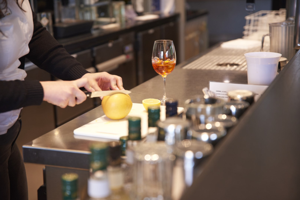 Person schneidet in einer Bar eine Grapefruit auf einem Schneidebrett, im Vordergrund sind ein Cocktail und Flaschen zu sehen.
