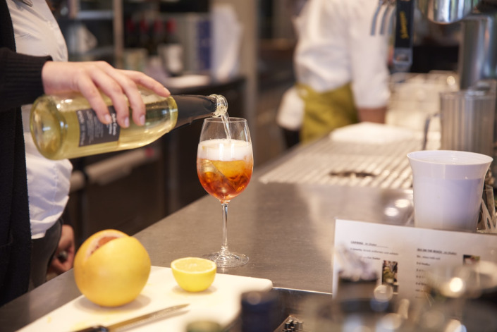 Eine Person gießt Weißwein in ein Glas mit Eis auf einer Bartheke, neben einer Grapefruit, einer Zitrone und einer Cocktailkarte.