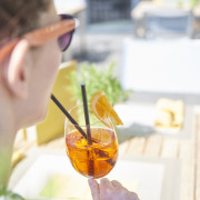 Eine Person mit Sonnenbrille hält an einem Tisch im Freien einen Orangencocktail mit Strohhalm und einer Orangenscheibe als Garnitur.
