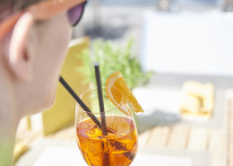 Eine Person mit Sonnenbrille hält an einem Tisch im Freien einen Orangencocktail mit Strohhalm und einer Orangenscheibe als Garnitur.