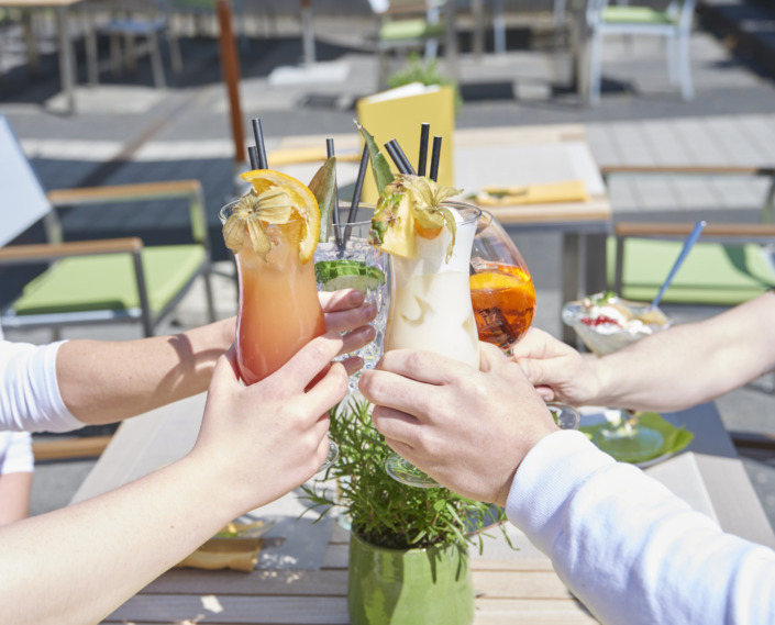 Drei Personen stoßen an einem sonnigen Tag an einem Tisch im Freien mit bunten, garnierten Cocktails an.
