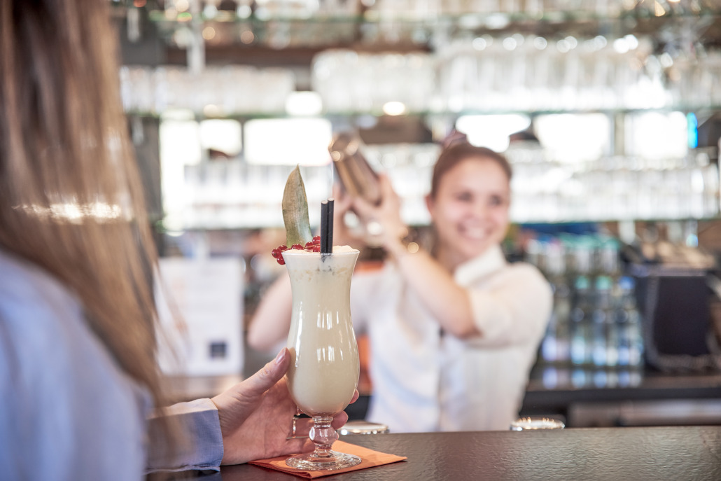 Eine Person hält an einer Bar einen tropischen Cocktail, während im Hintergrund ein Barkeeper einen Cocktailshaker schüttelt und lächelt.