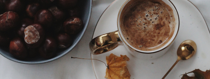 Eine gemütliche Herbstszene mit einer Tasse Kaffee, weißen Kürbissen, Kastanien, Herbstblättern und einem Wollschal auf einem weißen Tuch.