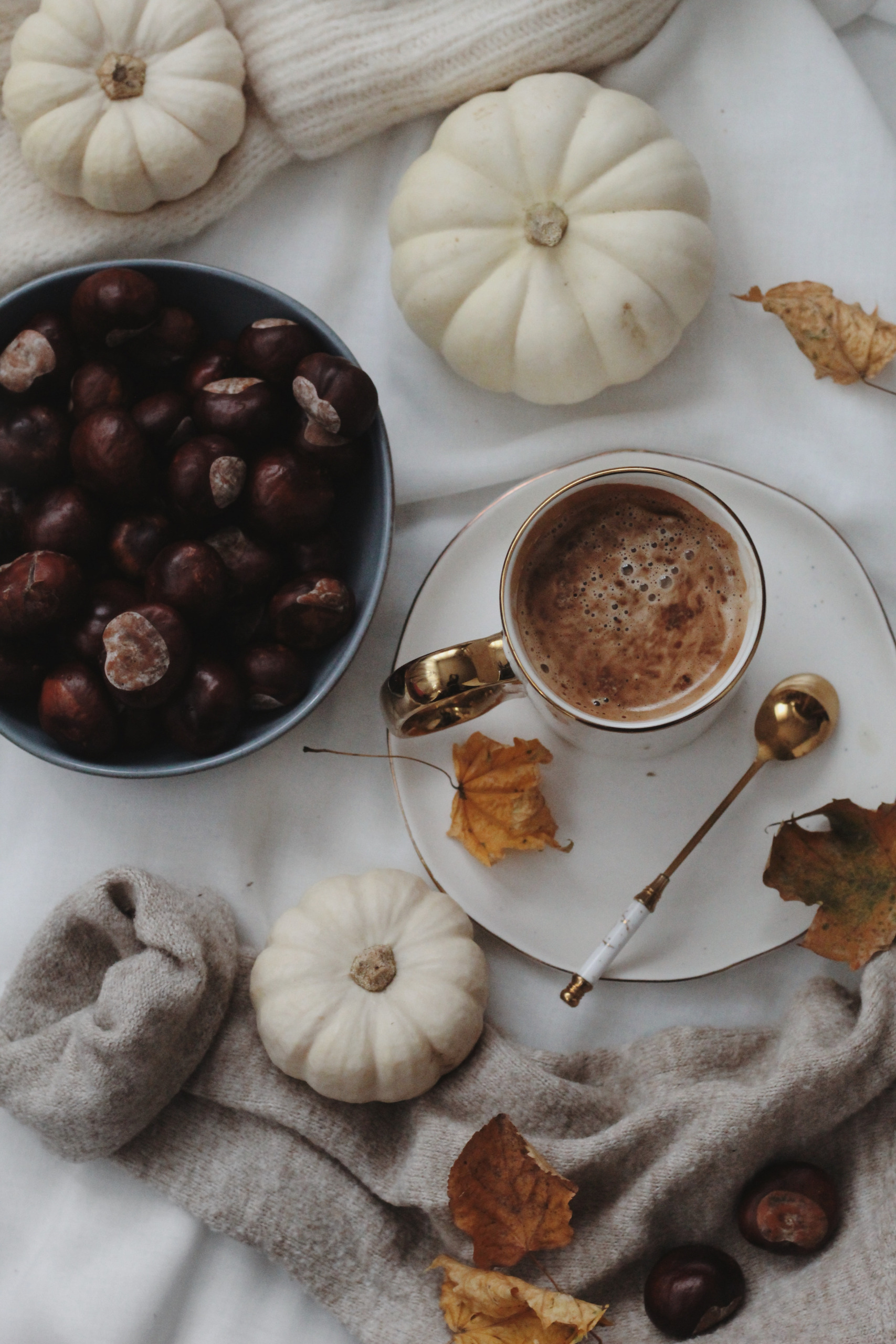 Eine gemütliche Herbstszene mit einer Tasse Kaffee, weißen Kürbissen, Kastanien, Herbstblättern und einem Wollschal auf einem weißen Tuch.
